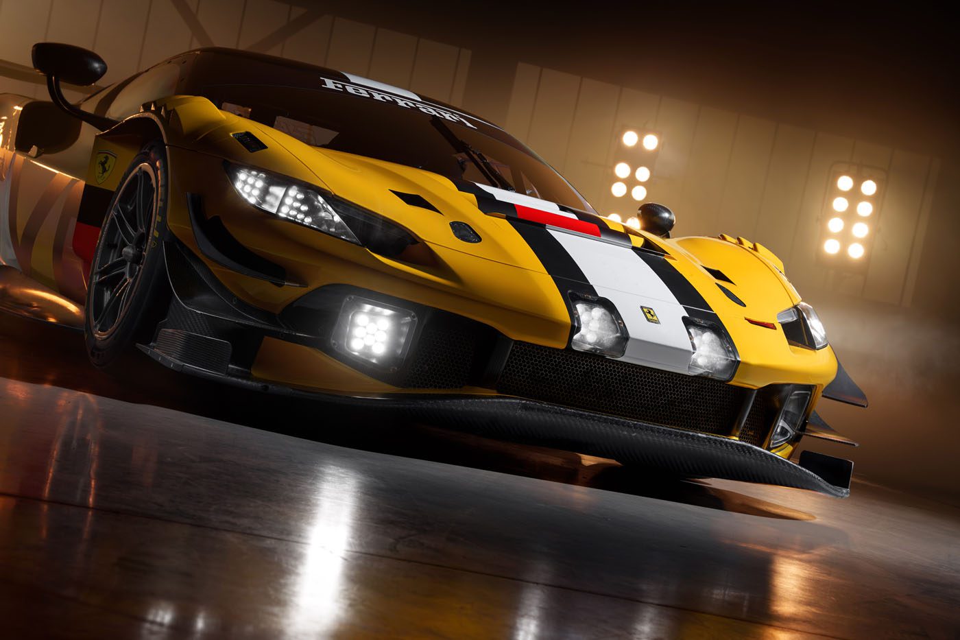 A yellow Ferrari 296 GT3 Evo race car with black and white striping is parked indoors under bright lights, viewed from a low front angle, capturing the spirit of the 24 Hours Of Spa.