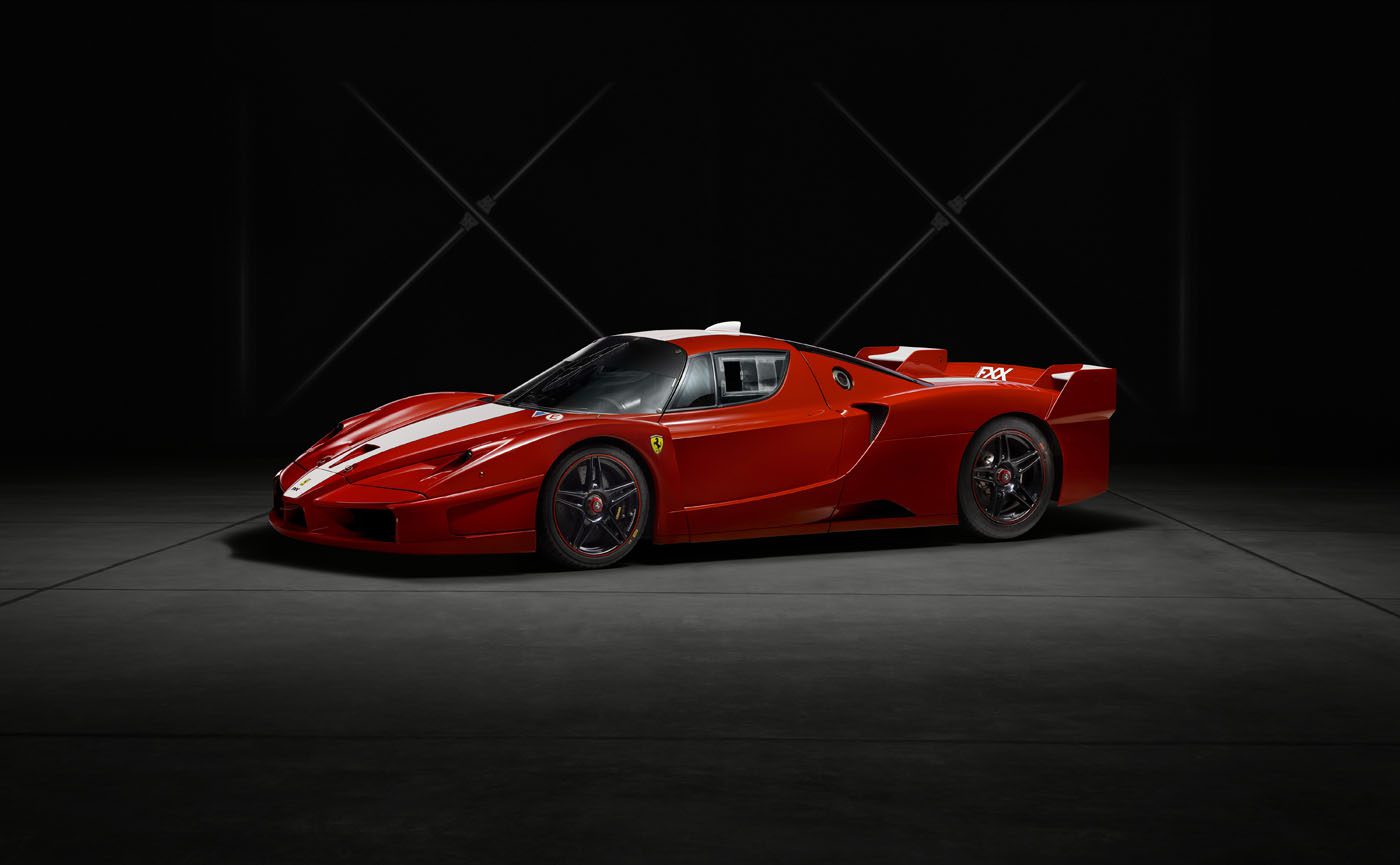 A red Ferrari FXX sports car from the exclusive XX Program is parked indoors on a dark concrete floor, illuminated against a black background with minimal lighting.