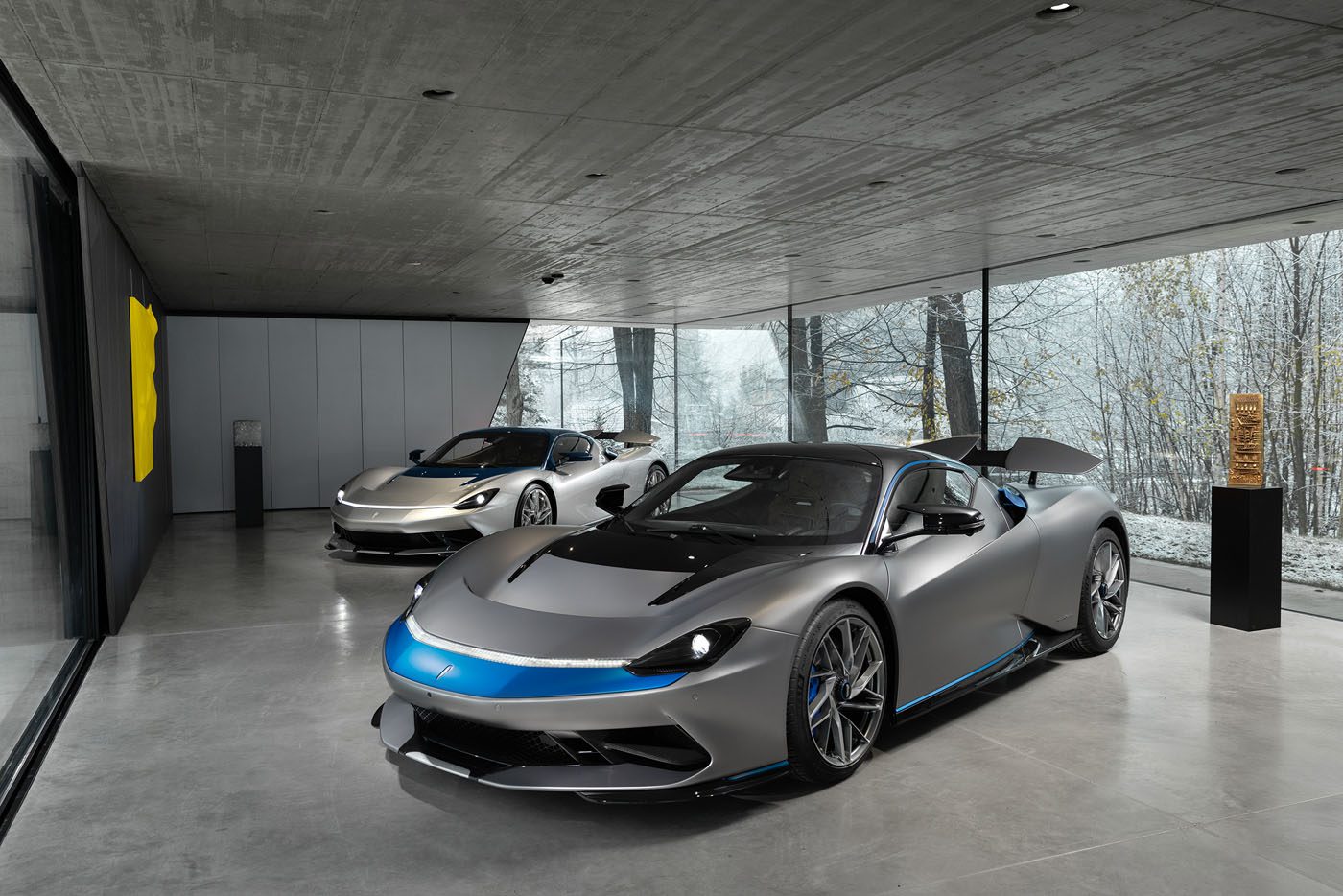 Two groundbreaking 2024 Automobili Pininfarina sports cars sit in a modern showroom with large glass windows, showcasing snowy trees outside. The cars flaunt a sleek silver and black color scheme highlighted by elegant blue accents.