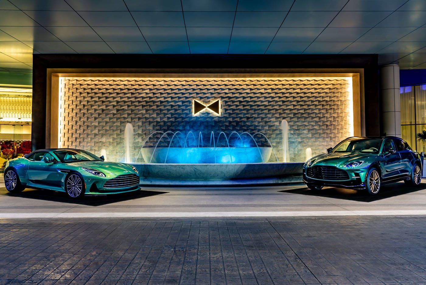 Two green Aston Martin cars from an exquisite car collection are parked in front of a large, illuminated fountain, capturing the vibrant spirit of Las Vegas at night.