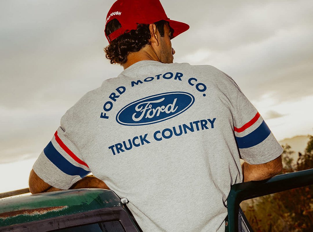 A person wearing a red cap and a gray Ford Motor Co. "Truck Country" shirt from the Limited Edition Collection leans on a green vehicle, viewed from behind.