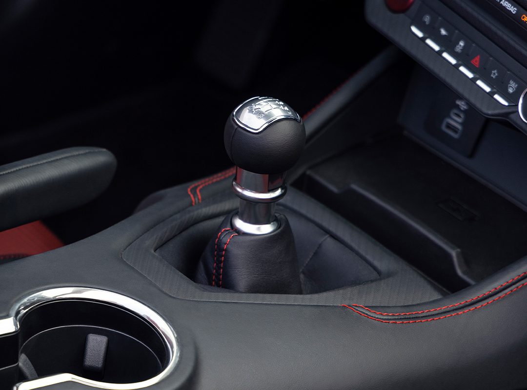 A manual transmission gear shift in a Ford car interior features striking red stitching. The dashboard controls and convenient cup holder add to the sleek design.