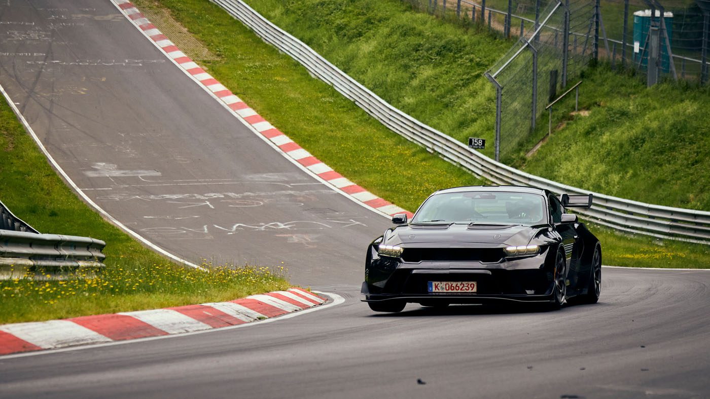 A black Ford Mustang GTD tears through the curvy, hilly racetrack of Nürburgring, surrounded by lush greenery.