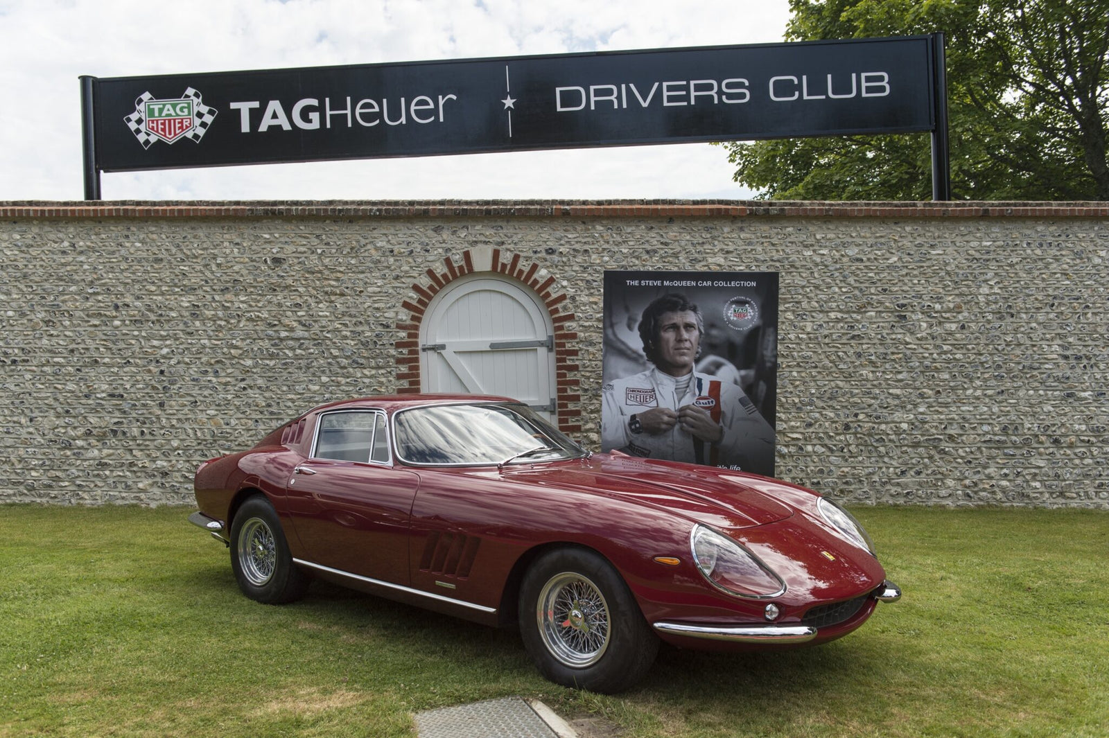 A classic red Ferrari sports car is parked on grass in front of a stone wall with TAG Heuer Drivers Club signage and a large racing driver poster at the Goodwood Festival of Speed, where TAG Heuer is the Official Timing Partner.