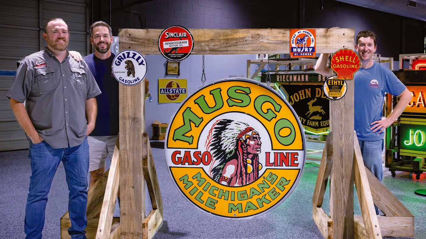 Three men stand beside a large vintage Musgo Gasoline sign, surrounded by collectible signs in an indoor setting—a testament to the enduring investment appeal of retro gas station memorabilia.