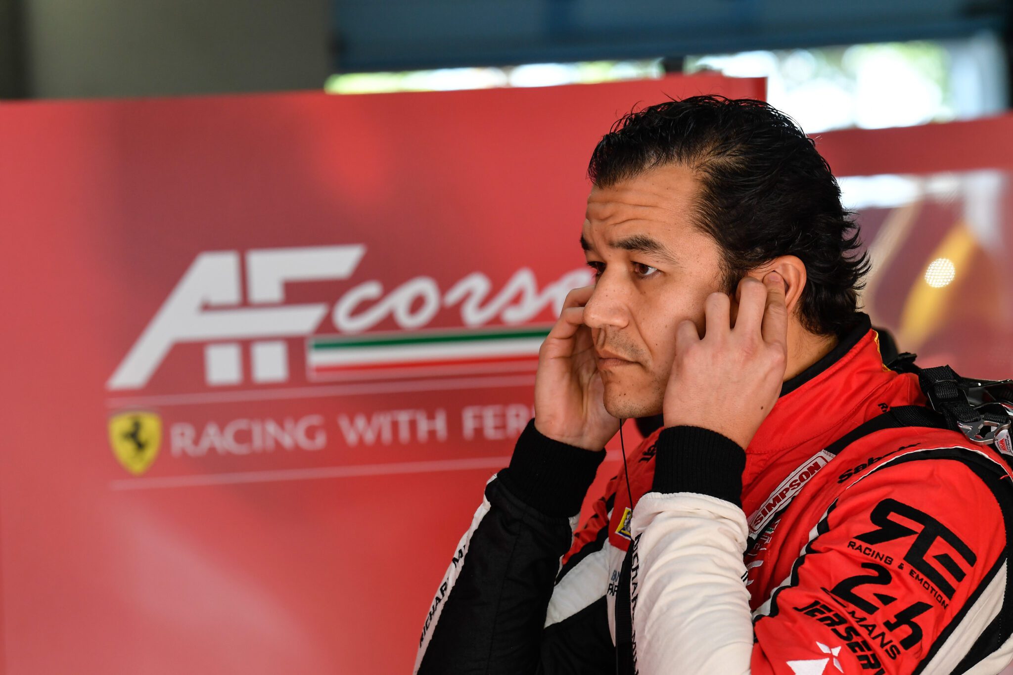 A race car driver in a red racing suit stands in front of an AF Corse Ferrari sign, covering his ears with his hands, as the duPont REGISTRY Group brings luxury and excitement to the racing world.