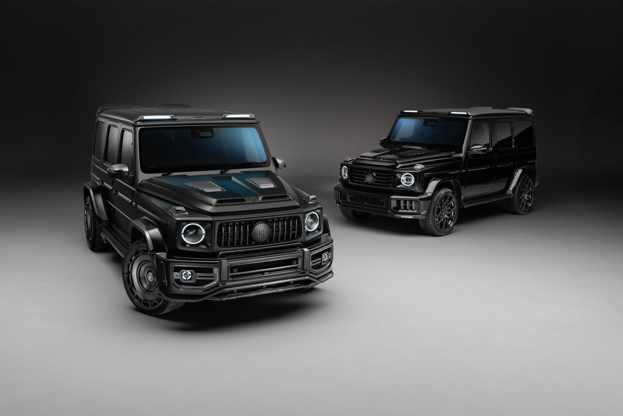 Two black luxury SUVs, including a G-Wagon with Widetrack & Aero Kits, are parked on a smooth, neutral-colored surface in an Urban Automotive-inspired studio setting with soft, even lighting.