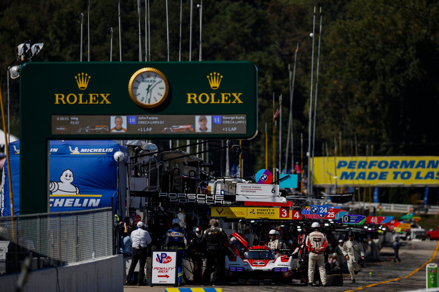Racing teams and cars are stationed in the pit lane under a large Rolex clock and signage at Petit Le Mans, capturing the excitement of the motorsport event.