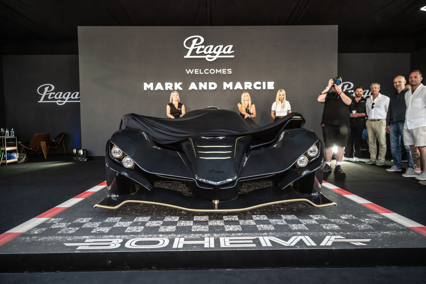 A black Praga Bohema supercar covered by a cloth is displayed on a stage, with a "Praga" sign and "Welcome Mark and Marcie" message in the background. Several people stand around the car at this US event.