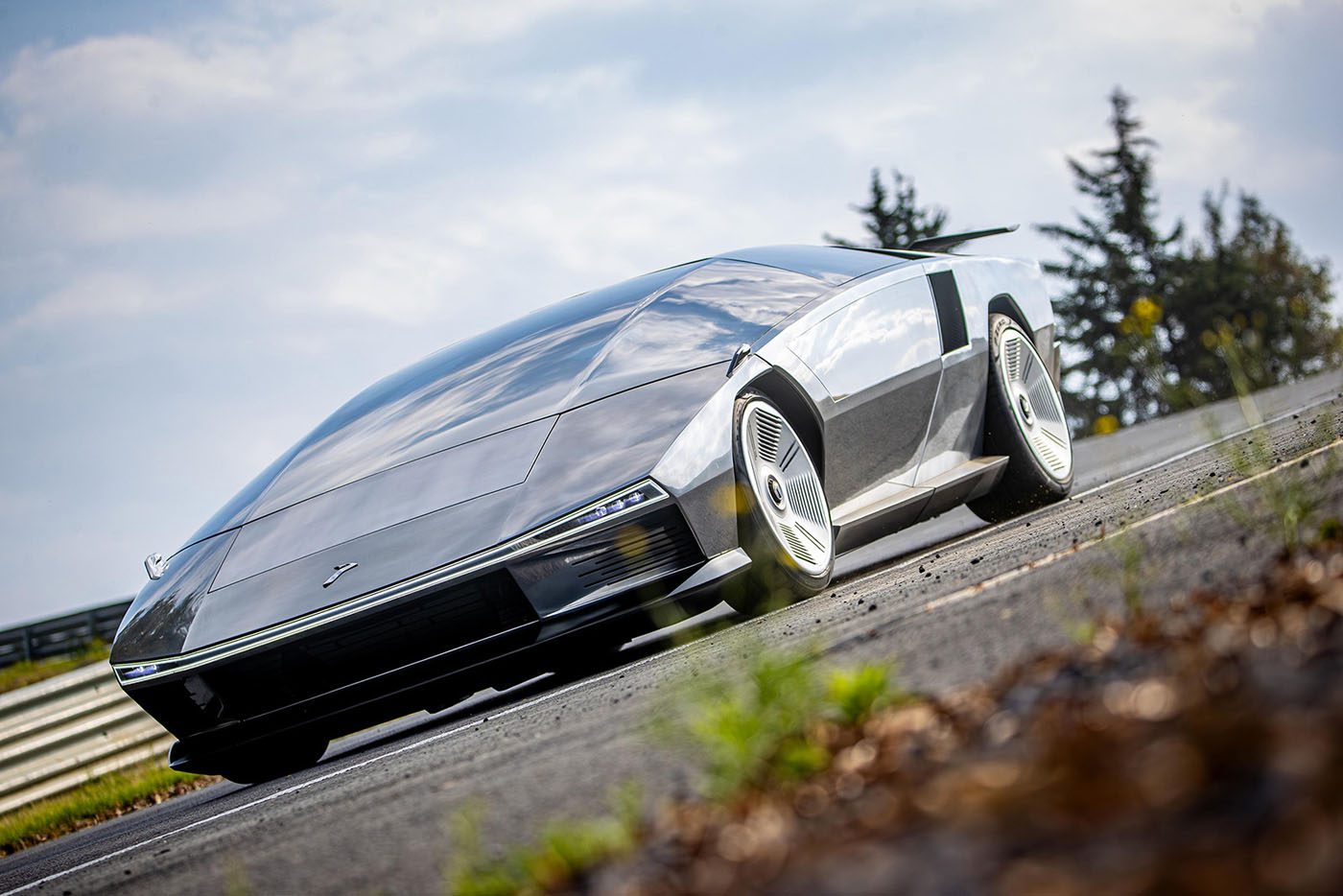 A futuristic, silver Peralta S supercar with a sleek, angular design is parked on a road with grass and trees in the background, evoking the innovation seen at events like the Monterey Motorsports Festival.