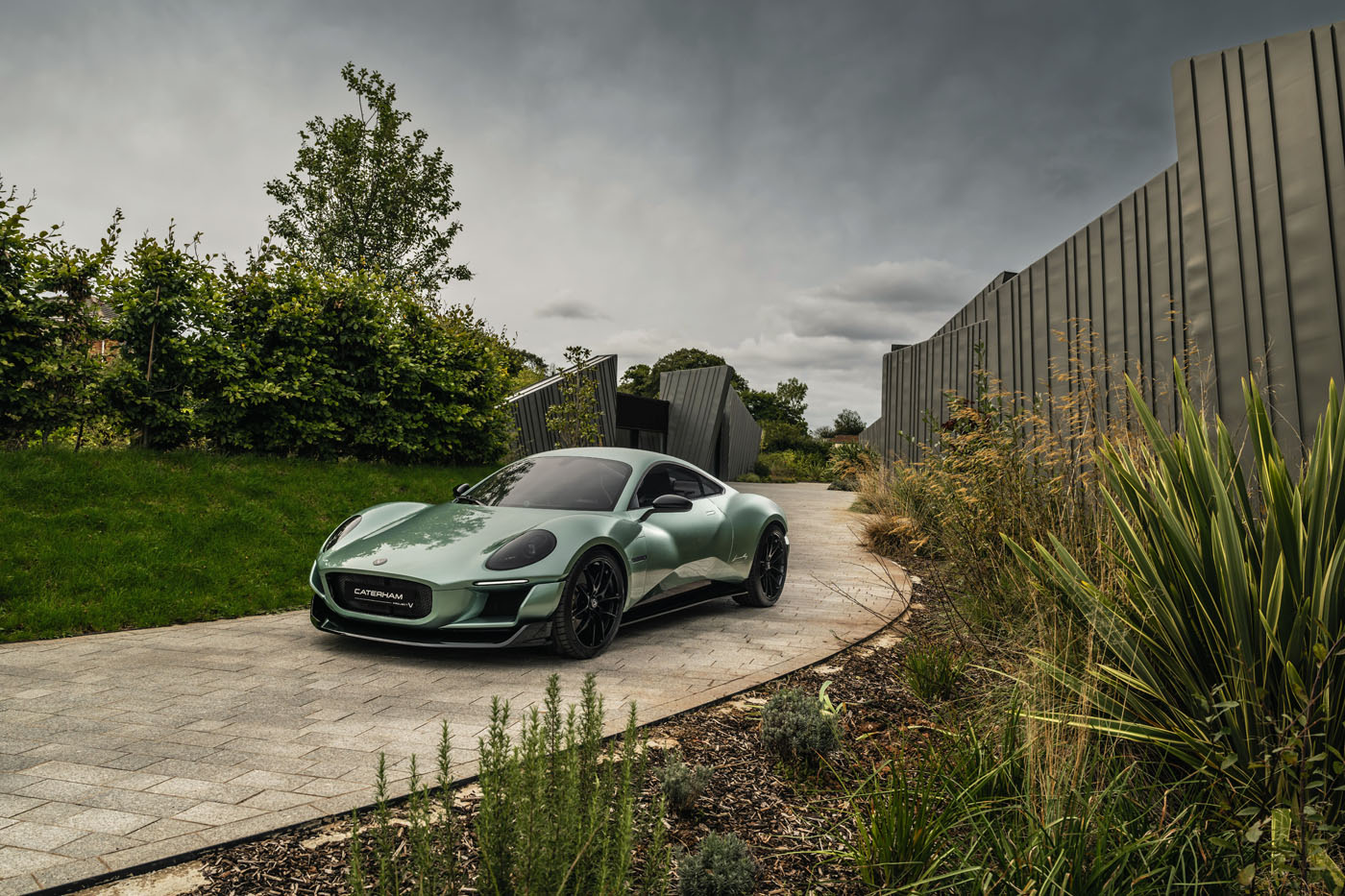A green sports car is parked on a paved driveway beside modern fencing and landscaping under a cloudy sky.