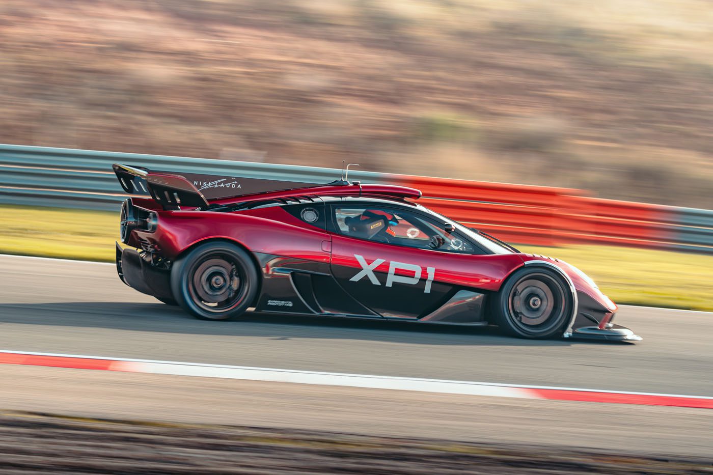A red sports car labeled "XP1" from Gordon Murray Special Vehicles speeds down a race track with blurred background and safety barriers, capturing the excitement of its debut at Monterey Car Week.