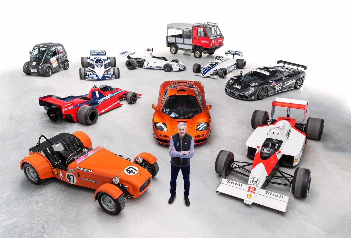 A person stands in front of a diverse lineup of vehicles, featuring race cars, a sports car, a microcar, and a small truck—each a testament to innovative automotive design—all displayed on a pristine white background.
