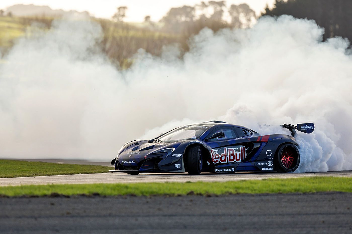 A black Red Bull Drift Car drifts on a track at the Monterey Motorsports Festival, producing a large cloud of white smoke from its rear tires.