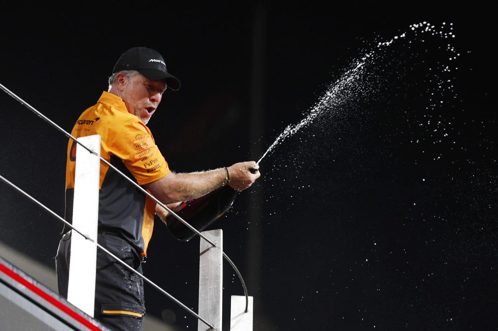 A jubilant person in team attire, perhaps celebrating a McLaren F1 victory, sprays champagne from a bottle on the platform.