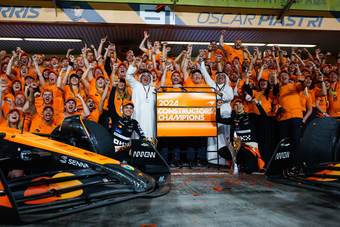 A large group with a World Championship Mindset celebrates an F1 championship win in front of a sign reading "2024 Constructors Champions" amid car parts and confetti.