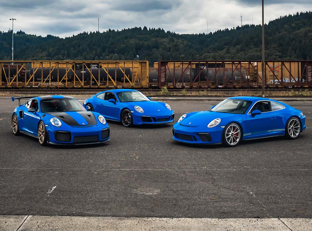 Three blue Porsche sports cars, including a Porsche 991 GT2 RS and a GT3 Touring, are parked on asphalt in front of a freight train with trees and hills in the background.