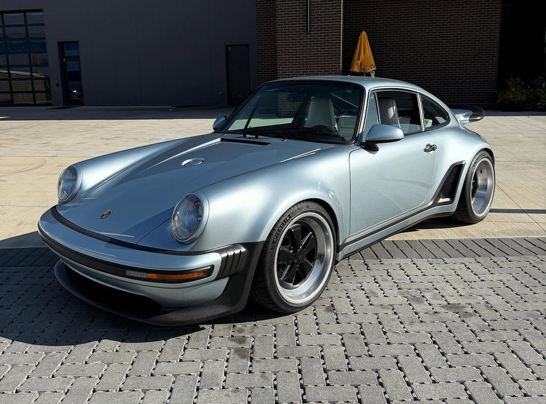 A silver classic Porsche sports car with black accents, part of the $3.8M Inventory at Graham Rahal Performance, is parked on a paved surface in front of a modern industrial building.