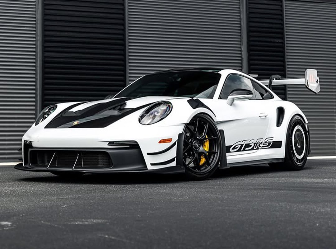 A white Porsche 911 GT3 RS sports car with black accents and a large rear wing is parked in front of a modern, striped metal wall.