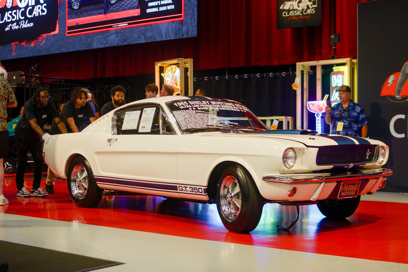 A white 1965 Shelby Mustang GT350 with blue racing stripes is displayed indoors at the GAA Classic Cars Greensboro NC Auction, surrounded by people and illuminated by event lighting.