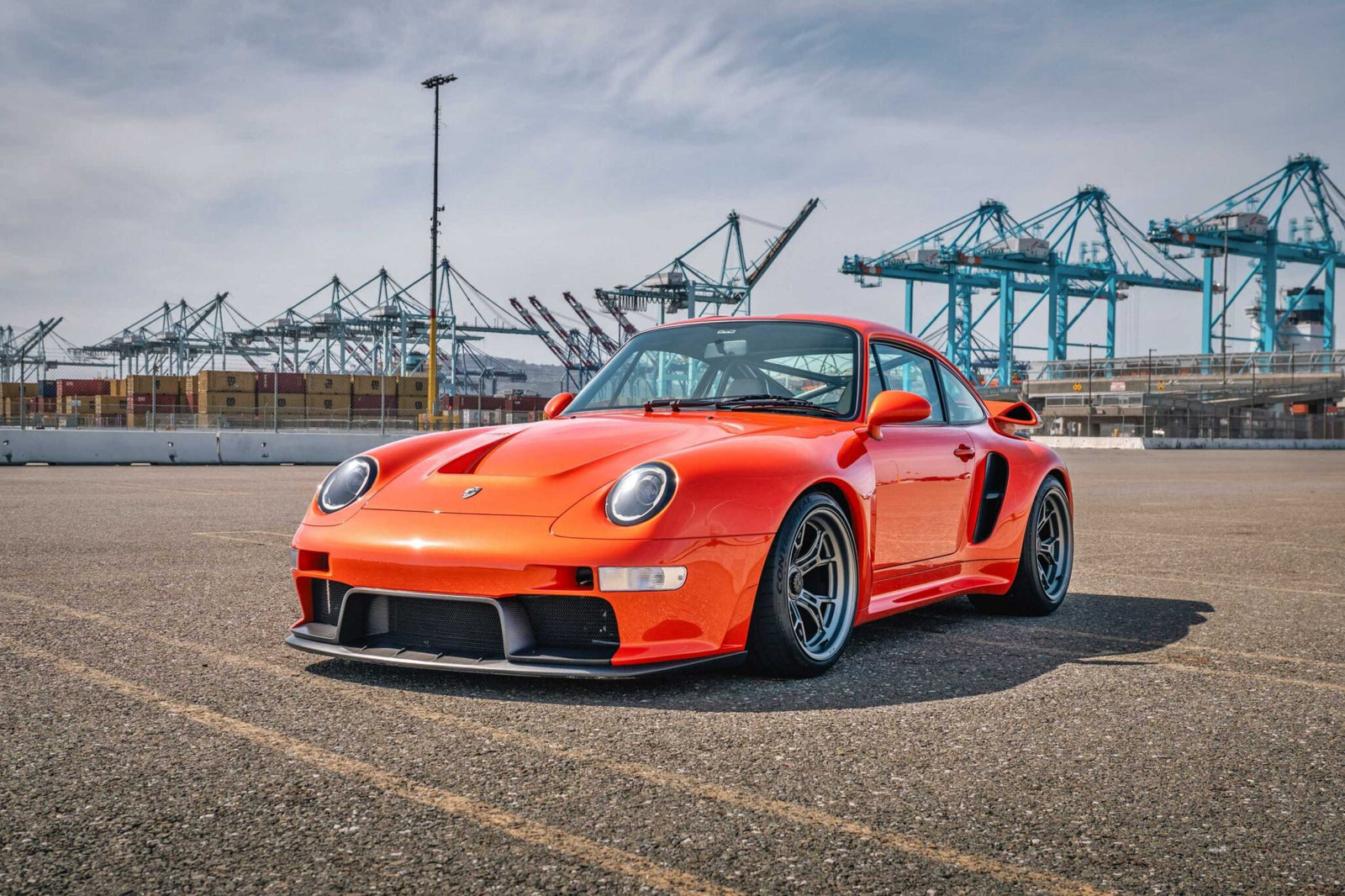 An orange Gunther Werks Turbo Tested sports car is parked on asphalt near a port with shipping containers and cranes in the background under a partly cloudy sky—bar none, the gnarliest Porsche restomod yet.