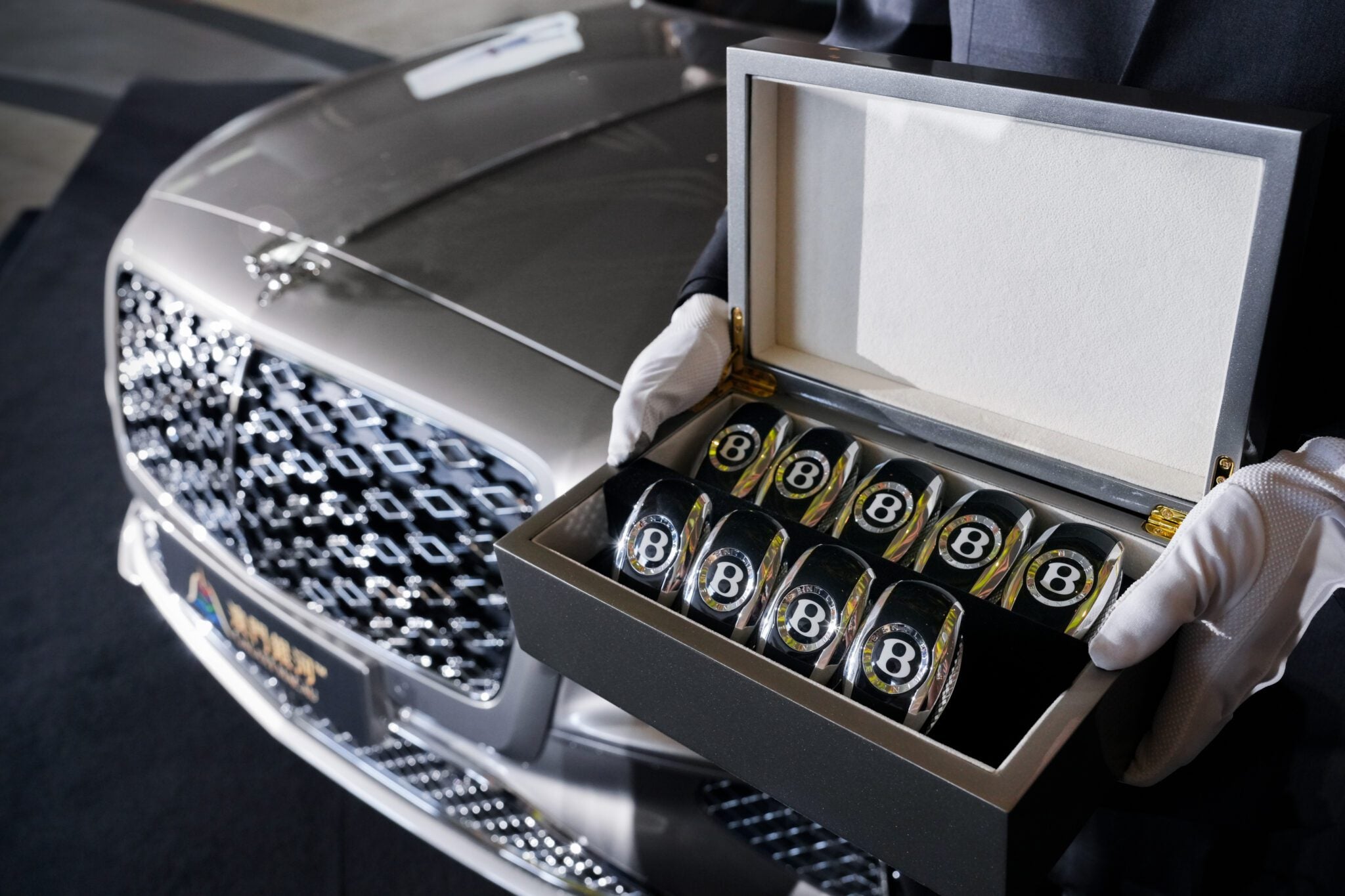 A person wearing white gloves holds an open box containing several Bentley-branded glass tumblers in front of a parked Bentley Motors Bespoke Sedan.