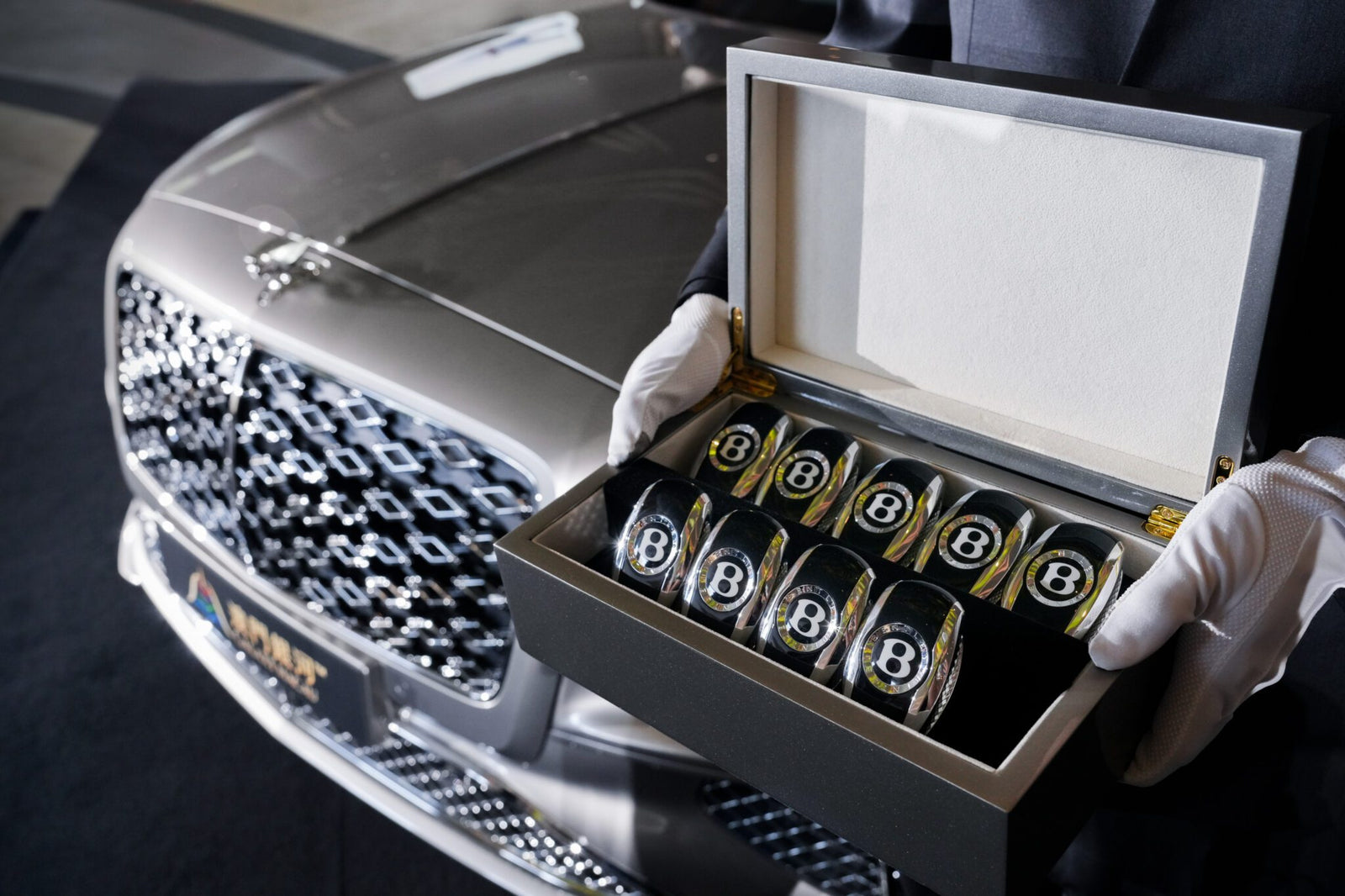 A person wearing white gloves holds an open box containing several Bentley-branded glass tumblers in front of a parked Bentley Motors Bespoke Sedan.