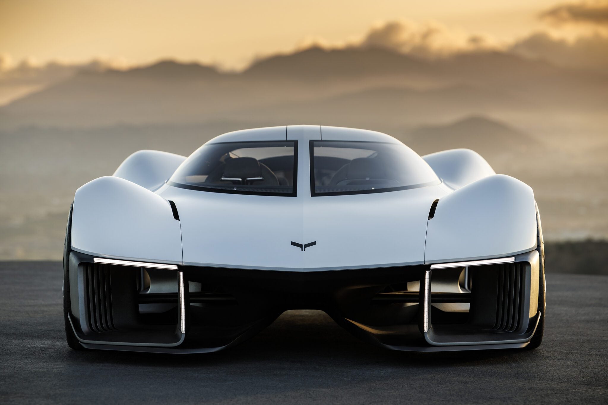 Futuristic white sports car with sleek design, wide stance, and distinctive front grill from a renowned UK Design Studio, set against a sunset backdrop with mountains.