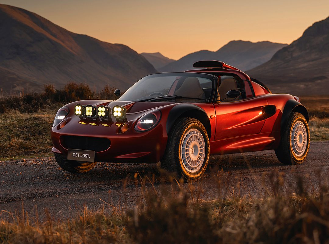 A red sports car with off-road tires and extra lights on the hood is parked on a rural road at sunset. The license plate reads "GET LOST." Mountains are visible in the background.