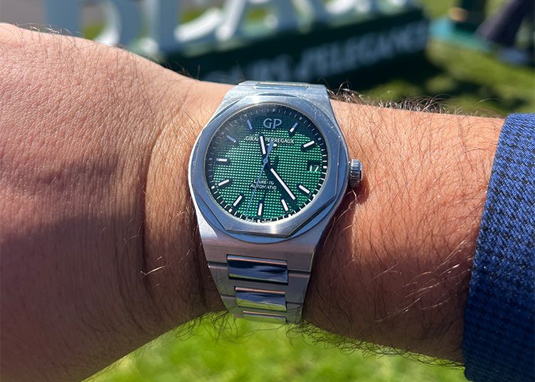 A Girard-Perregaux stainless steel wristwatch with a green textured dial and silver hour markers is worn on a person's wrist outdoors—perfect for making Monterey Car Week memories, reminding you that it's always time well spent.
