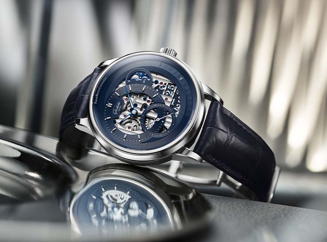 A silver luxury wristwatch by Glashütte Original, featuring a blue skeleton dial and black leather strap, is displayed on a metallic surface with a blurred background—an exquisite limited edition from the PanoMaticCalendar collection.