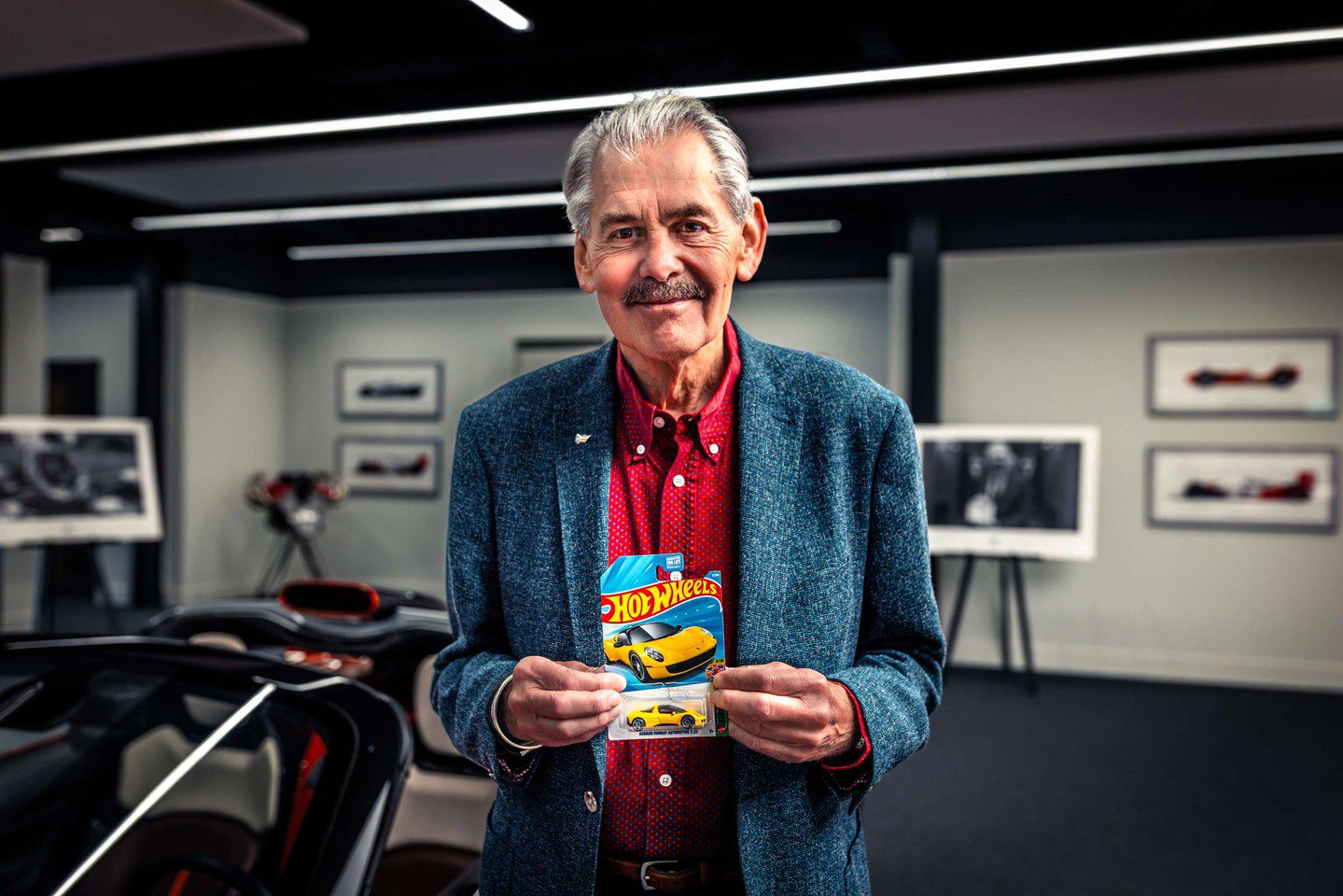 An older man in a blue blazer and red shirt holds a yellow Hot Wheels T.33 package in a room with framed photos, cars, and a nod to Gordon Murray in the background.