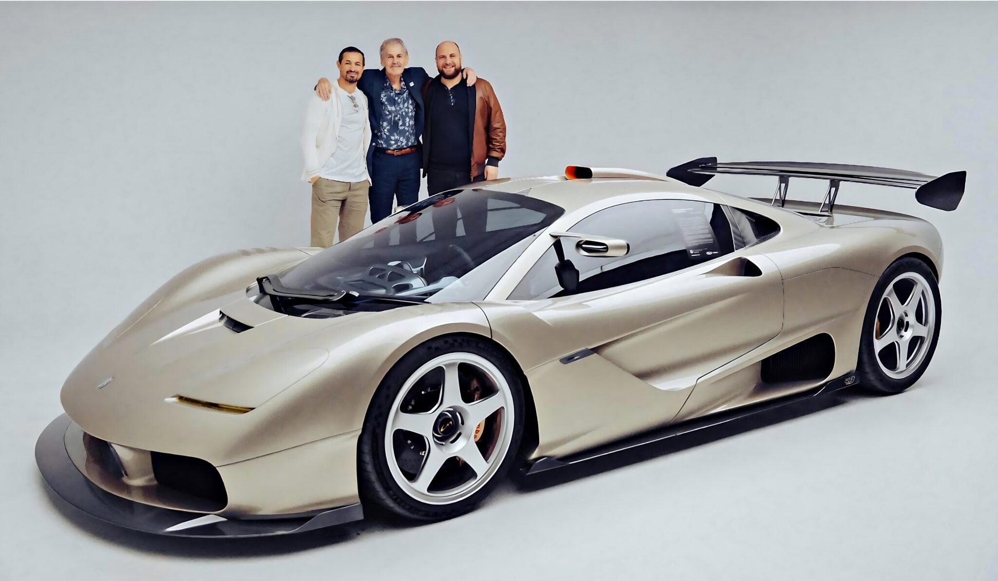 Three people stand behind a silver sports car with a large rear wing, photographed in a studio with a plain background, representing the innovative spirit of Gordon Murray Automotive.