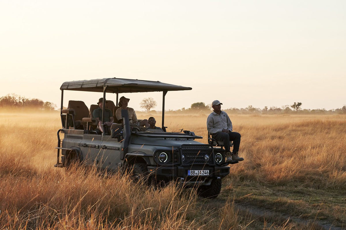 A group of people ride in an open INEOS Grenadier Game Viewer through tall, dry grass at sunset, with one person seated on the front of the vehicle.