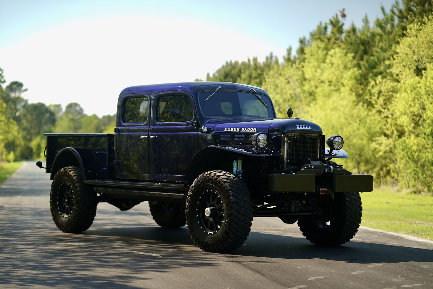 A restored 1947 Desert Power Wagon with large off-road tires is parked on a paved road, surrounded by greenery and trees—a striking tribute to classic trucks enhanced with Turbo Diesel power.