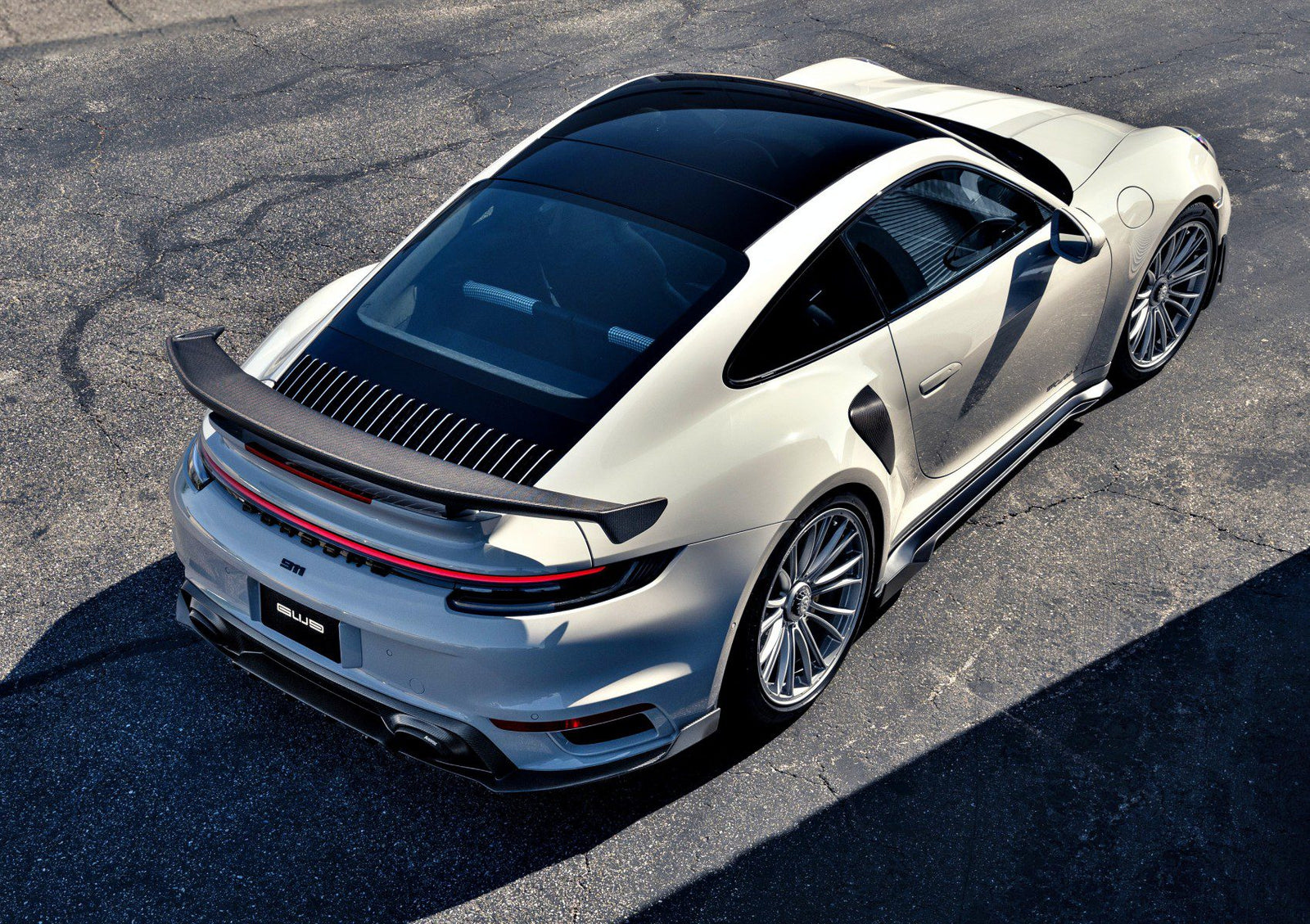 A silver sports car with a large rear spoiler, inspired by Gunther Werks Goes Modern: Meet GW9’s Carbon-Clad 992 Turbo S, is parked on an asphalt surface and viewed from above at an angle.