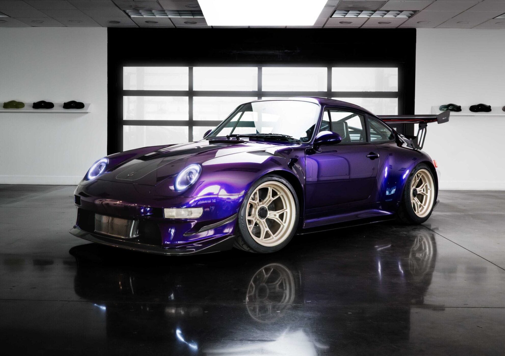 A remastering of the Porsche 911 by Gunther Werks, this purple sports car with a large rear wing is parked indoors on a polished concrete floor, with helmets displayed on shelves in the background.