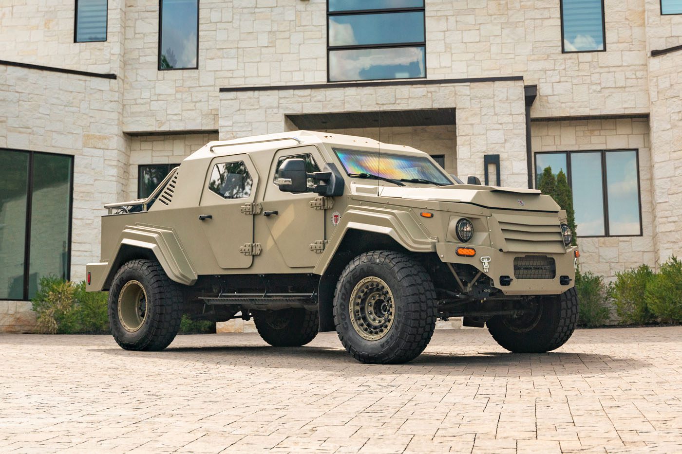 A tan 2020 Gurkha, a street-legal armored vehicle, is parked on a stone driveway in front of a modern light-colored building with large windows.