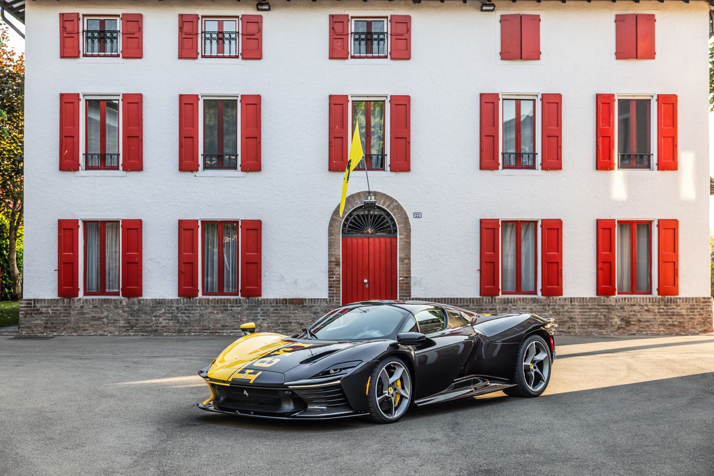 A black and yellow Tailor Made Daytona SP3 Ferrari sports car is parked in front of a white house with red shutters and a red door, capturing the excitement of Car Week Charity Auction.