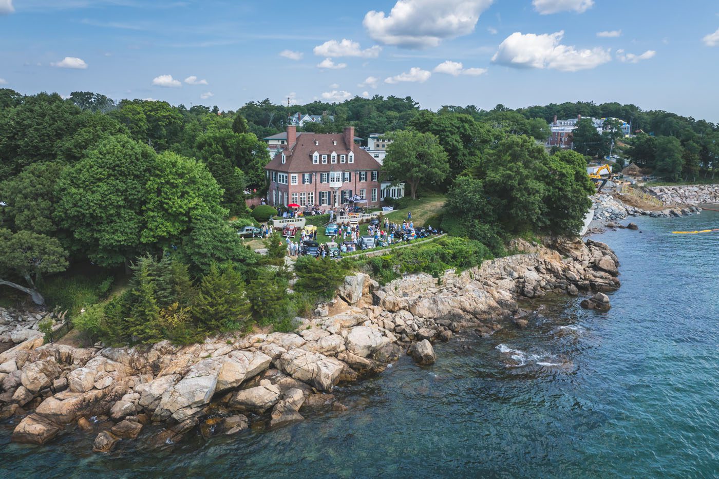 A large house on a rocky shoreline surrounded by trees, with people gathered on the lawn for the Misselwood Concours d’Elegance 15th Anniversary in July and the sea in the foreground under a partly cloudy sky.