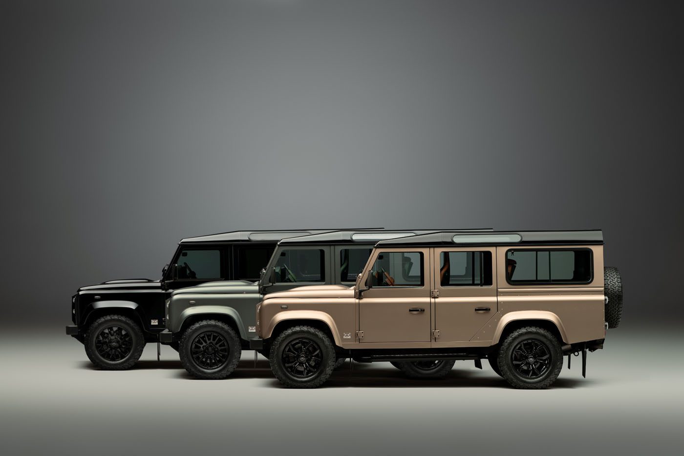 Three Land Rover Defender vehicles—including a Classic Defender and a Defender OCTA—in black, green, and beige are parked side by side in a studio setting with a neutral gray background.