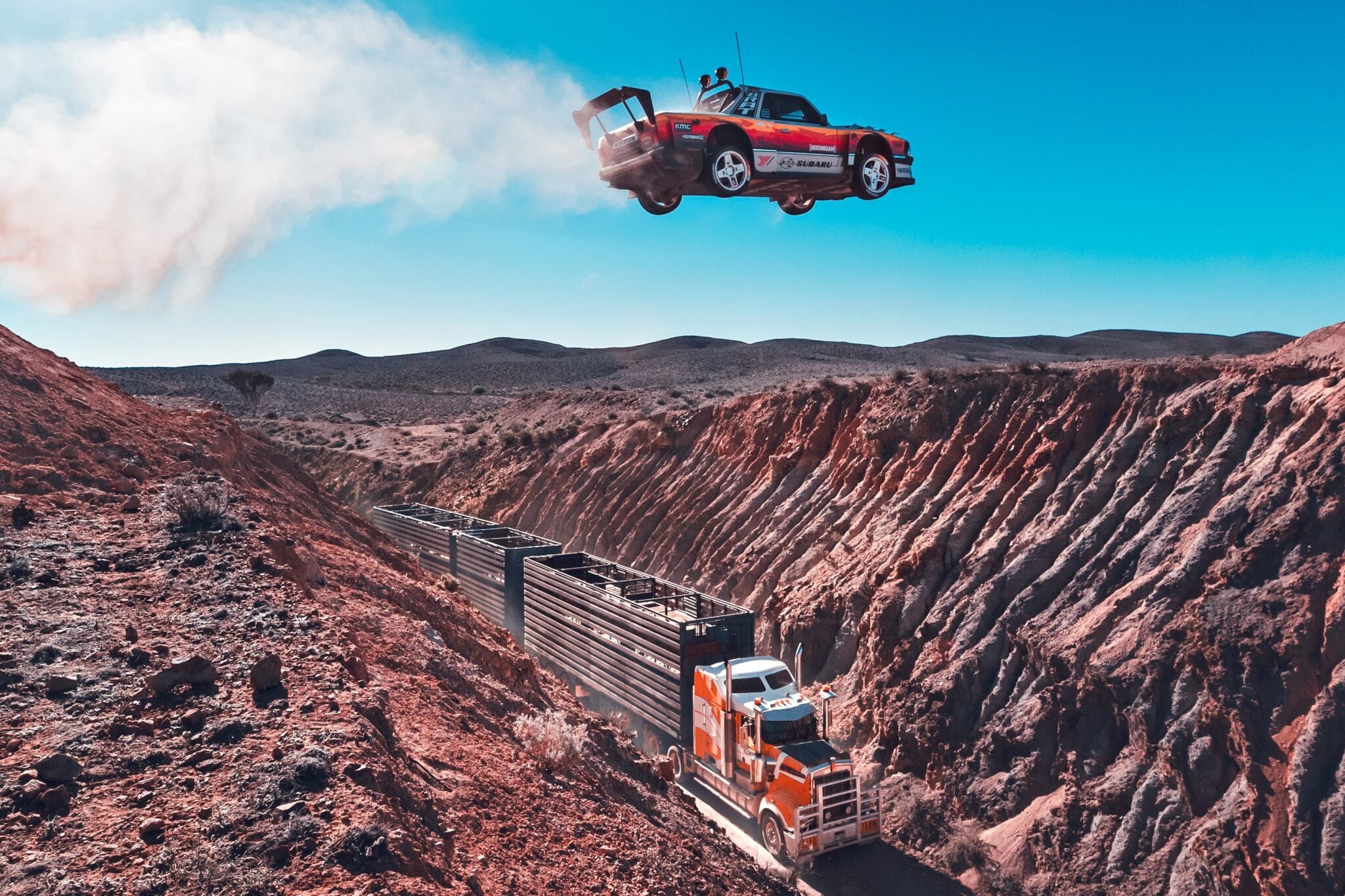 A red sports car launches off a ramp, soaring over a canyon and a moving semi-truck with trailers on a dirt road below—classic Pastrana Gymkhana Film action set in a breathtaking desert landscape.