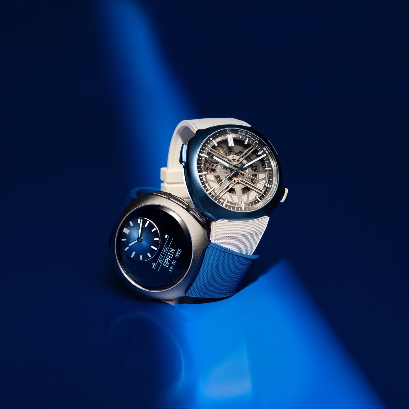 Two modern wristwatches, including a sleek H. Moser & Cie., are displayed on a blue surface—one with an analog face and white strap, the other digital with a silver-tone metal strap—both illuminated under a focused beam of light.