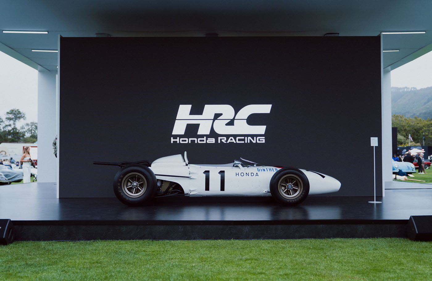 A vintage white Honda racing car with the number 11 is displayed on stage at The Quail 2025, set against a black backdrop featuring the Honda Racing Corporation logo—an elegant moment in the Unveiling Icons series.
