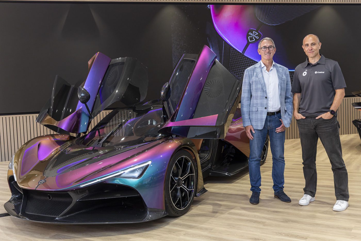 Two men stand next to a Zenvo sports car with open butterfly doors in a modern showroom, ready for Goodwood.