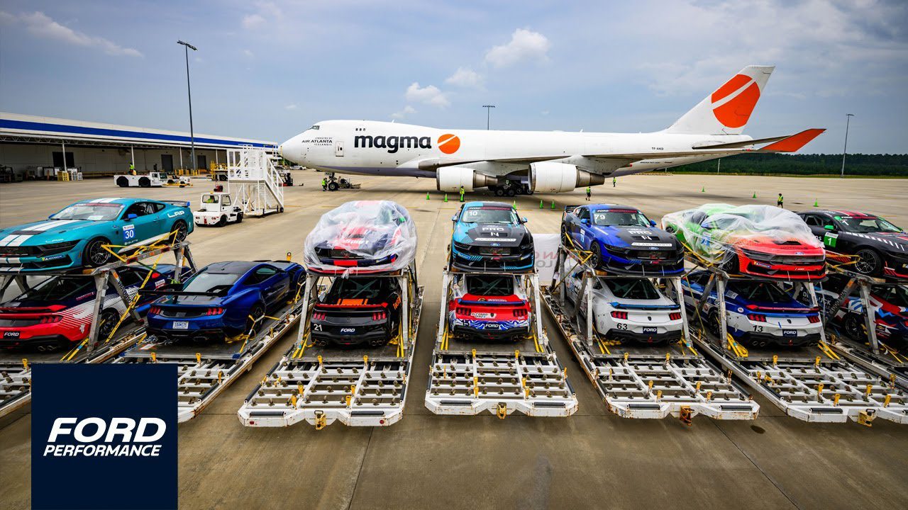 Multiple Ford sports cars, including the Mustang Dark Horse from Ford Performance, are secured on pallets in front of a Magma cargo airplane at an airport, ready for transport.