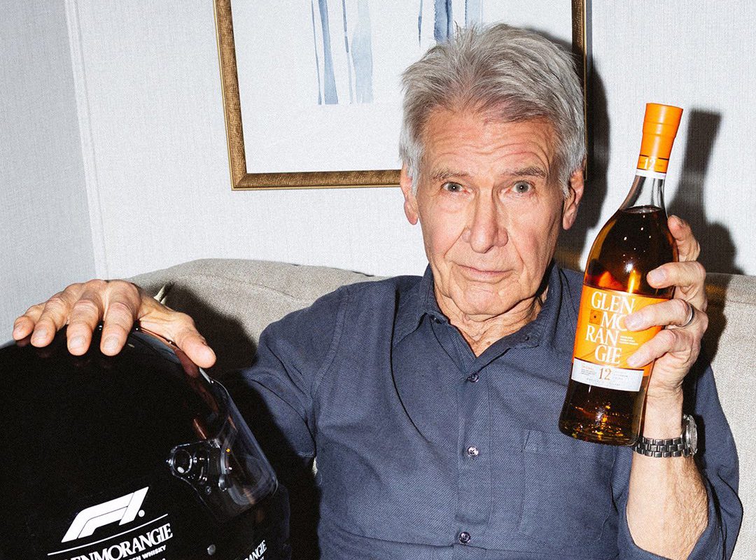 Older man with gray hair sits on a couch, holding a bottle of Glenmorangie whisky in one hand and a sleek Formula 1 racing helmet in the other.