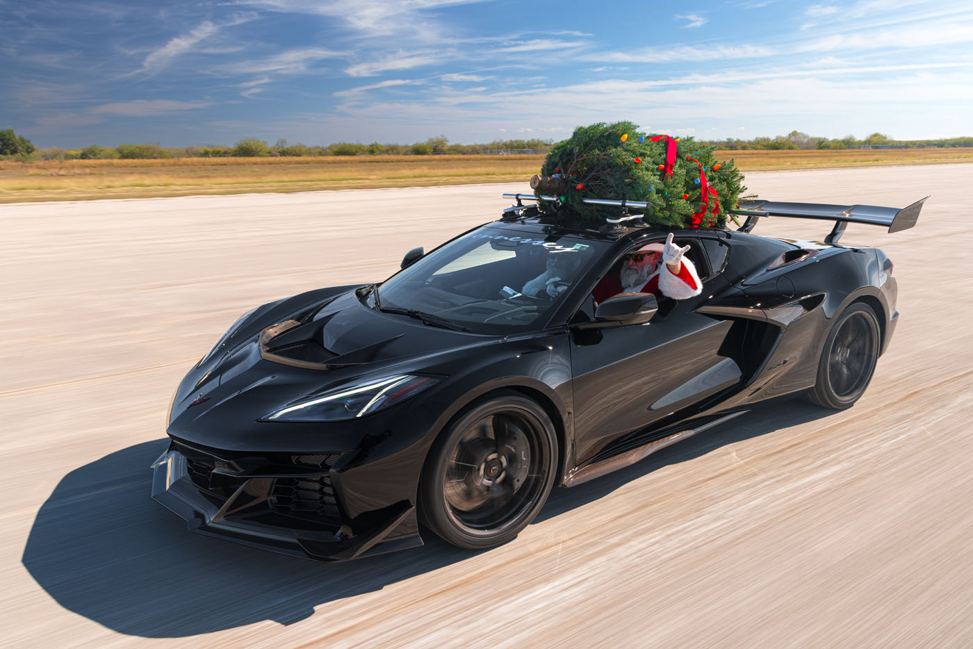 A person dressed as Santa Claus drives a 196-MPH Hennessey sports car with a Christmas tree secured on the roof, showcasing holiday spirit down an empty road.