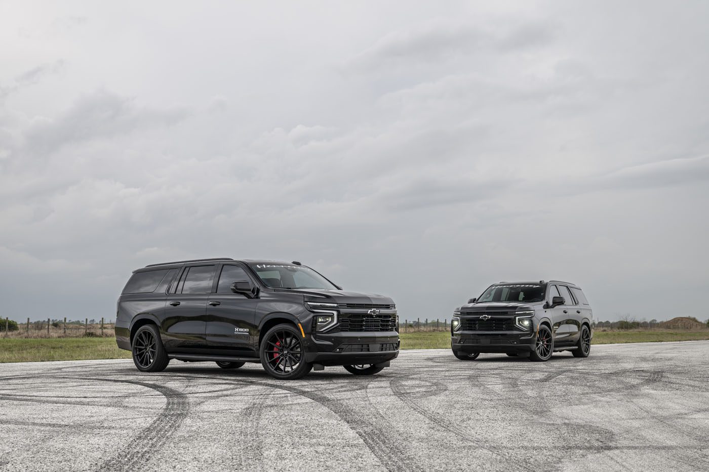 Two black SUVs, including a Hennessey-tuned Chevrolet Tahoe, are parked on an open pavement under a cloudy sky, grassy fields stretching in the background—a true Suburban Upgrade.