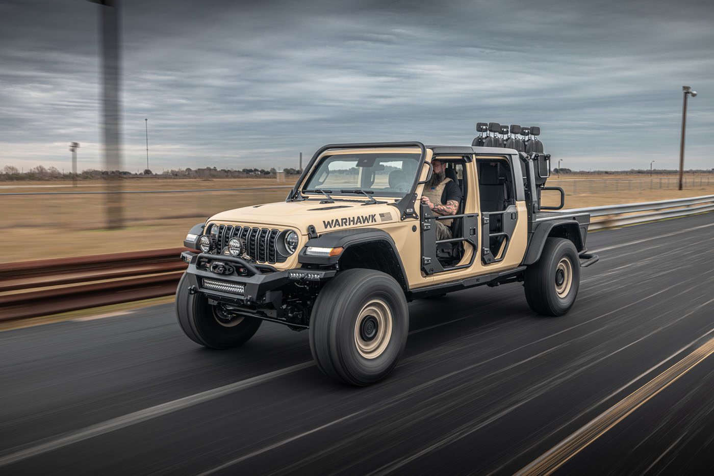 A beige modified off-road vehicle with large tires and no doors drives on a road, proudly labeled "WARHAWK" by Hennessey Defense Systems. A person is visible in the driver's seat.